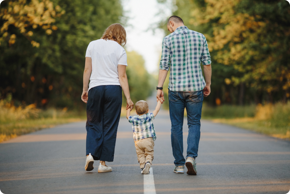 Family walking on path - parents and toddler holding hands from behind, father in plaid shirt and mother in white shirt walking with young child in matching plaid during golden hour, representing life transitions and family support during OCD treatment